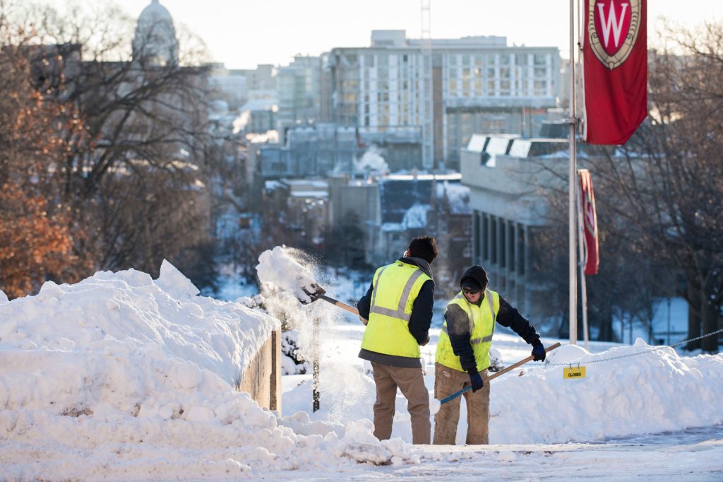 workers clearing snow with shovels on Bascom Hill as a "W" crest UW-Madison flag is above them along with the Wiconsin Capitol building in the distance