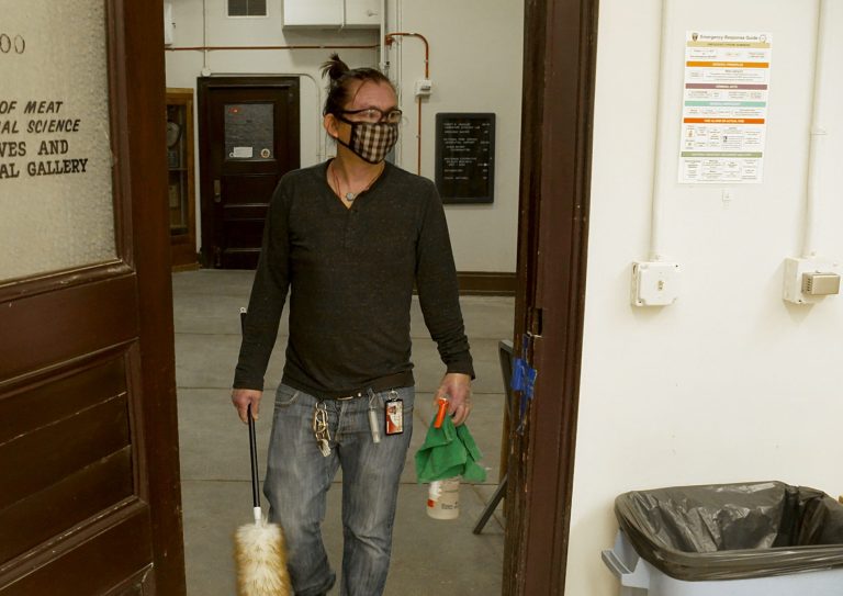 custodial worker entering a door while wearing a face covering, long sleeve dark shirt and jeans; while carrying cleaning solution, a rag, and microfiber duster. A lined trash canister is seen to the right of the door frame with colorful safety signage above it on the wall, alongside a light switch and wiring box